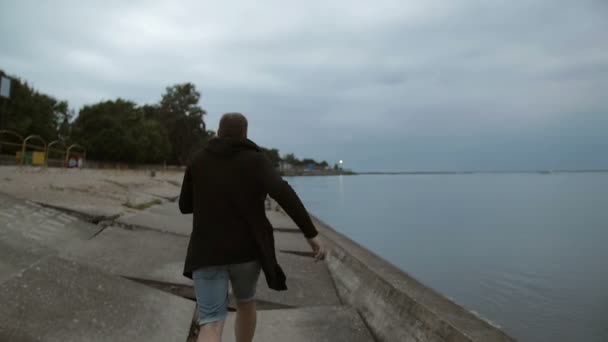 Mann im langen Pullover läuft am Strand entlang. Wasser, Bäume, Mond im Hintergrund, Zeitlupe, Rückansicht, Steadicam-Aufnahme — Stockvideo