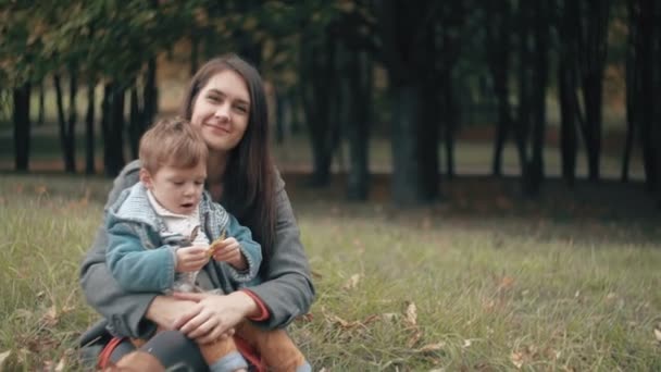 Young beautiful mother playing with her cute little son, who is sitting on her lap in the autumn park 4k — Stockvideo