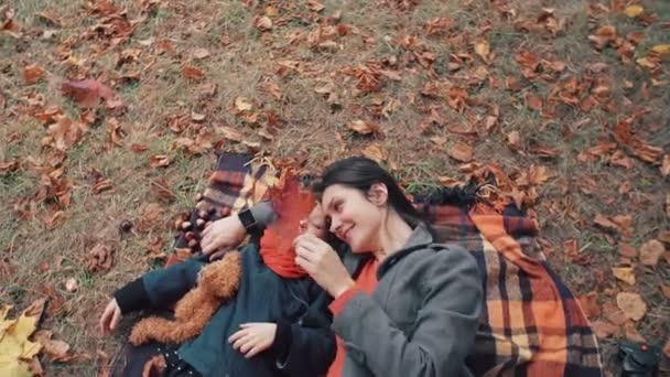 Mooie moeder met haar schattig dochtertje liggend op de plaid spelen met esdoorn bladeren in de herfst park 4k — Stockvideo