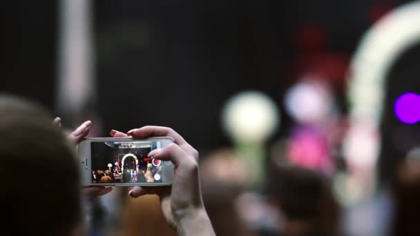 Primer plano de las manos sosteniendo un teléfono inteligente y tomando un video de un desfile de moda. Los modelos se pueden ver en la pantalla del teléfono inteligente — Vídeos de Stock