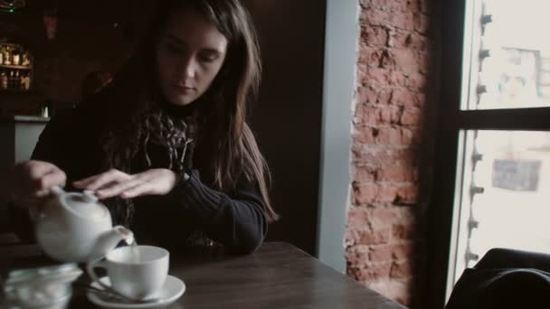 Chica joven vierte té de una tetera en una taza sentada en la cafetería moderna — Vídeo de stock