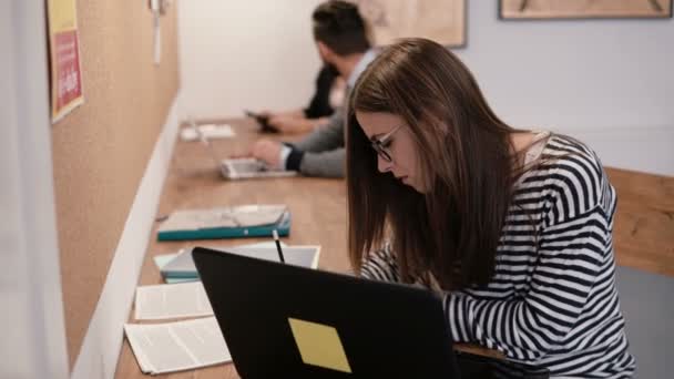 Menina atraente jovem está usando um laptop, trabalhando com documentos, fazendo anotações no escritório de inicialização moderna — Vídeo de Stock