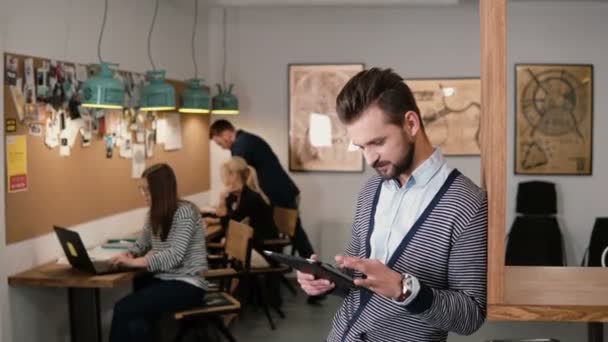 Joven hombre barbudo guapo utiliza tableta de pantalla táctil en la moderna oficina de inicio . — Vídeos de Stock