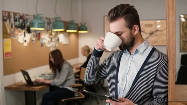 Jeune homme barbu beau utilise tablette écran tactile et boire du café dans le bureau de démarrage moderne . — Video