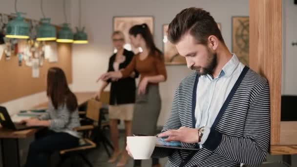 Jeune homme barbu beau utilise tablette écran tactile et boire du café dans le bureau de démarrage moderne . — Video