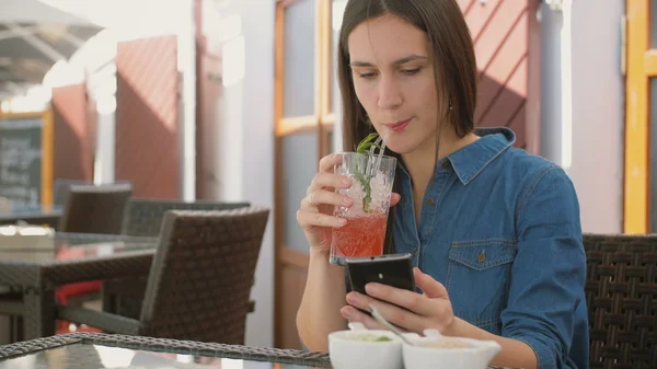 Брюнетка пользуется смартфоном, сидя в кафе, выпивая и наслаждаясь прохладным напитком из соломенной трубки. 4k , — стоковое фото