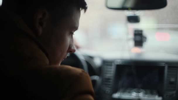 Un homme assis dans une voiture, regardant en bas et en haut. La main gauche est sur le volant. Des gouttes de pluie sur la fenêtre avant. Vue arrière — Video