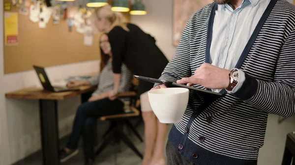 Крупный план рук молодой человек использует планшет с сенсорным экраном и пьет кофе в современном офисе стартапа . — стоковое фото