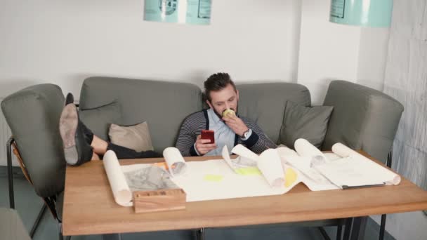 Jonge man rust het vermijden van het werk zittend op de Bank gebruik smartphone en eten apple zet zijn voeten op tafel — Stockvideo