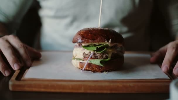 Sabrosa hamburguesa es grande en una bandeja de madera. Un hombre toma sus manos y come, prueba el sabor — Vídeos de Stock