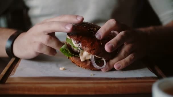 Sabrosa hamburguesa es grande en una bandeja de madera. Un hombre toma sus manos y come, prueba el sabor — Vídeo de stock