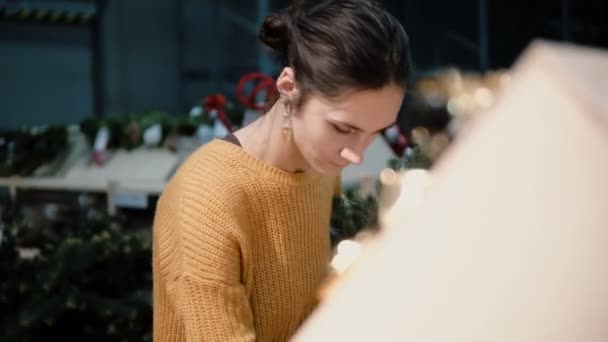 Joven chica morena atractiva en la tienda elige un árbol de Navidad artificial y juguetes, decoración de Navidad . — Vídeo de stock