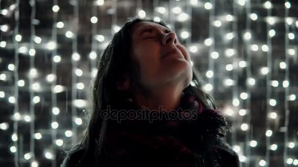 Mujer atractiva joven disfrutando de la nieve que cae en la noche de Navidad delante de la pared decorativa llena de luces brillantes — Vídeos de Stock