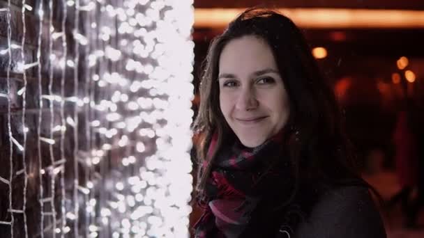 Junge attraktive Frau im fallenden Schnee in der Weihnachtsnacht, die auf die Kameraleuchten im Hintergrund blickt — Stockvideo