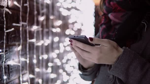 Närbild händer ung kvinna med smartphone i den fallande snön på jul natten stående nära lampor vägg, — Stockvideo