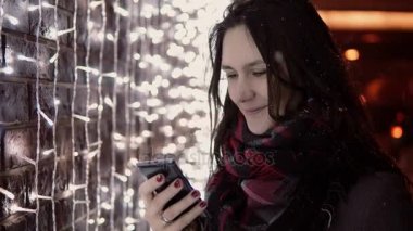 Noel gece ayakta ışıklar duvarının yakınında, düşen kar içinde smartphone kullanan genç çekici kadın,