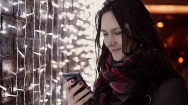 Fiatal, vonzó nő használ smartphone a hulló hó Karácsony este állandó fények fal közelében, — Stock videók