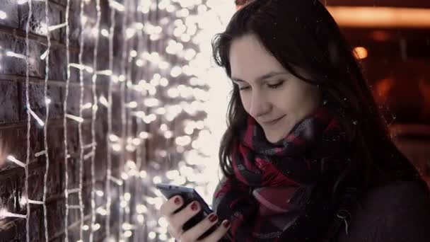 Jovem mulher atraente usando smartphone na neve caindo na noite de Natal de pé perto de luzes parede , — Vídeo de Stock