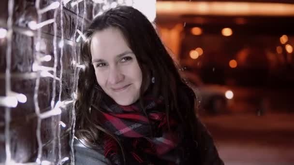 Young attractive woman at snowy Christmas night smiles looking at the camera standing near lights wall and walks away — Stock Video