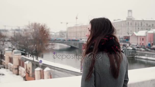 Vista traseira. Menina morena jovem atraente de pé na ponte e olha para a cidade de inverno coberta de neve . — Vídeo de Stock