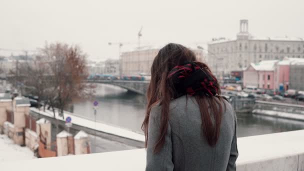 Vista trasera. La triste chica morena de pie en el puente suspira y mira a la ciudad de invierno cubierta de nieve . — Vídeos de Stock