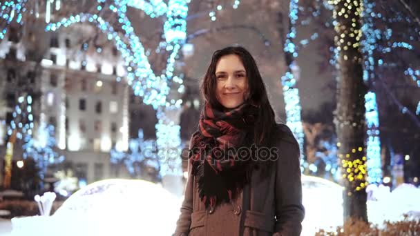 Приваблива жінка на засніженій різдвяній ніч посміхається, дивлячись на камеру перед парковими деревами, прикрашеними блискучими вогнями — стокове відео