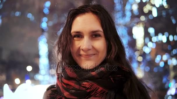 Attractive woman at snowy Christmas night smiles looking at the camera in front of park trees decorated sparkling lights — Stock Video