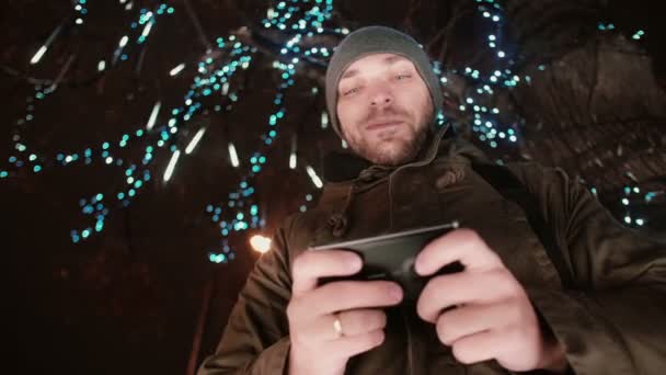 Young handsome man using smartphone at Christmas night standing under a tree decorated with sparkling lights — Stock Video