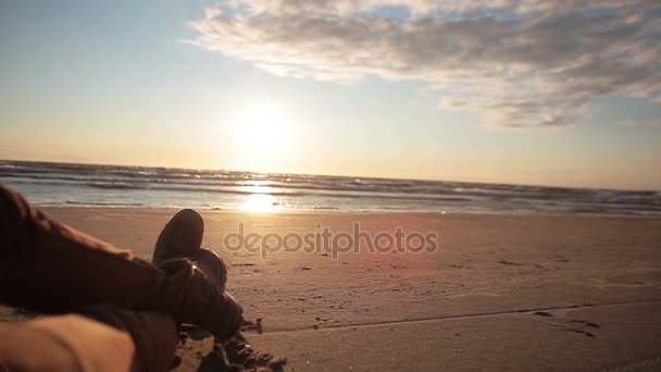 มุมมองของคนที่ข้ามขาขณะนอนบนชายหาดทรายเพลิดเพลินกับพระอาทิตย์ตกพาสเทลที่สวยงาม . — วีดีโอสต็อก