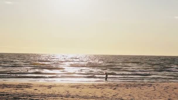 Una joven con un vestido blanco está caminando en una playa de arena iluminada por el sol con olas que llegan directamente a sus pies — Vídeos de Stock