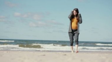 Hamile bir esmer genç kadın çıplak ayakla yavaş yavaş kameraya yaklaşırken kumlu bir plaj yürüyor.