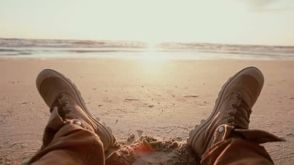 A close up van twee voeten in bruine laarzen rustend op een zandstrand bij dageraad. — Stockvideo