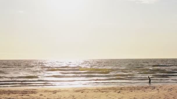 Uma vista distante de uma jovem que está de pé em uma praia de areia iluminada pelo sol olhando para ondas cobrindo seus pés . — Vídeo de Stock