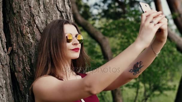 Beautiful young woman with long dark hair in sunglasses taking selfie, at the park at sunset. slow mo — Stock Video