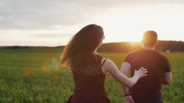 Szczęśliwy człowiek goni jej człowiek działa w dziedzinie o zachodzie słońca. Womans włosów kołysać się na wietrze. Od tyłu, slow mo, steadicam strzał — Wideo stockowe