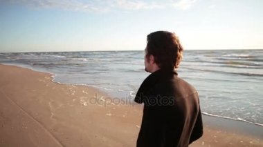 siyah montlu genç adam onun arkada parlayan güneş ile deniz kıyı yürüyor.