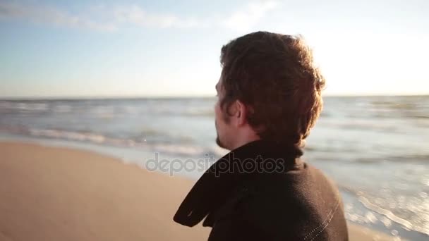 Una vista lateral de un joven moreno con un abrigo negro camina a lo largo de la orilla del mar . — Vídeos de Stock