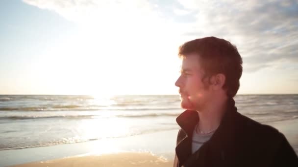 Una vista laterale di un giovane uomo dai capelli scuri con un cappotto nero che si trova sulla riva del mare godendo il tramonto — Video Stock