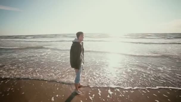 Dos chicos jóvenes están empapando sus pies en las olas del mar mirando a la playa y hablando . — Vídeo de stock