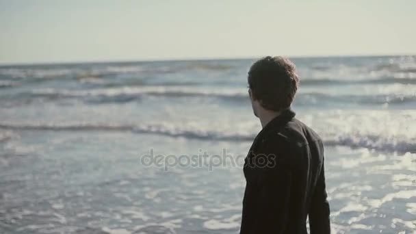 Um jovem está andando ao longo da bela praia em um dia ventoso carregando seus tênis em suas mãos . — Vídeo de Stock