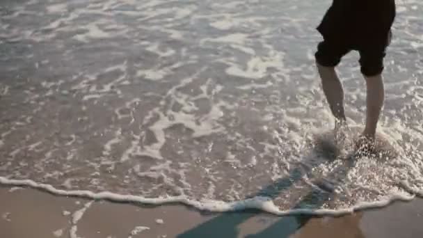 Un joven está caminando en una hermosa playa con sus pantalones vaqueros enrollados y empapando sus pies en el mar . — Vídeo de stock