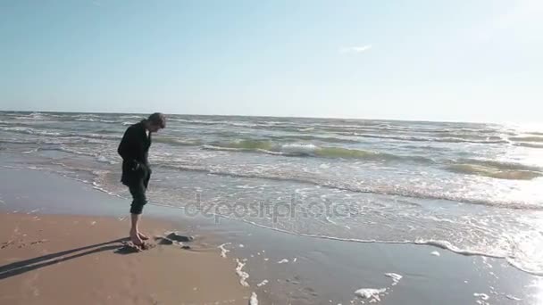 Молодий чоловік стоїть босоніж на красивому піщаному пляжі і копає дірку в піску пальцями . — стокове відео