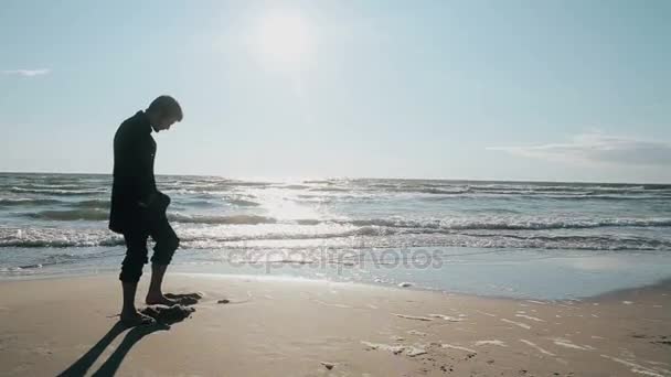 Sidovy av en ung man står barfota på stranden på en härlig solig dag gräver ett hål i sanden med tårna. — Stockvideo