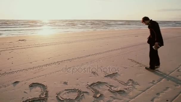 Ciemnowłosa facet, który ma na sobie czarny płaszcz i dżinsy jest pisanie nie b w piasku na plaży. — Wideo stockowe