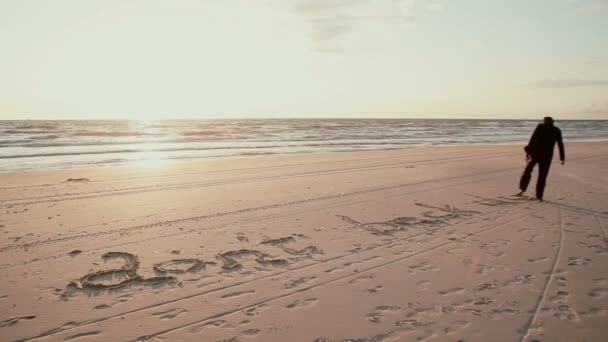 На закате молодой парень в черном пальто пишет на песчаном пляже: . — стоковое видео