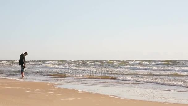 若い男が彼の足をカバーする波を見て海の水で彼の足を浸漬します。. — ストック動画