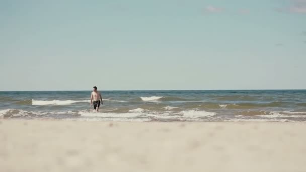 Un joven atlético de cabello oscuro con pantalones cortos negros está saliendo del mar sin camisa. . — Vídeo de stock