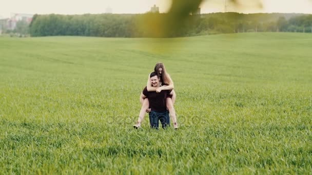 2 人の恋人間の幸せな瞬間。ブルネットの女性彼女の恋人の背中にジャンプの笑みを浮かべて、彼はフィールドに彼女を運ぶ。遅い mo — ストック動画