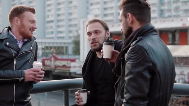 Tres jóvenes guapos con barba hablan, sonríen y toman café en el camino cerca de una barandilla de puente en la ciudad. Lento mo — Vídeos de Stock