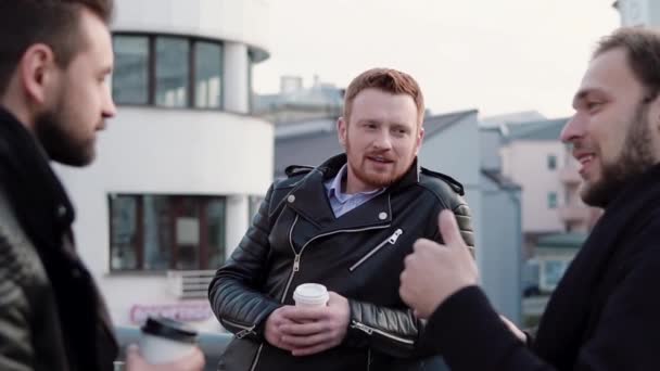 Trois beaux jeunes hommes barbus parlent, sourient et prennent un café en allant près d'une rampe de pont dans la ville. Moteur lent — Video